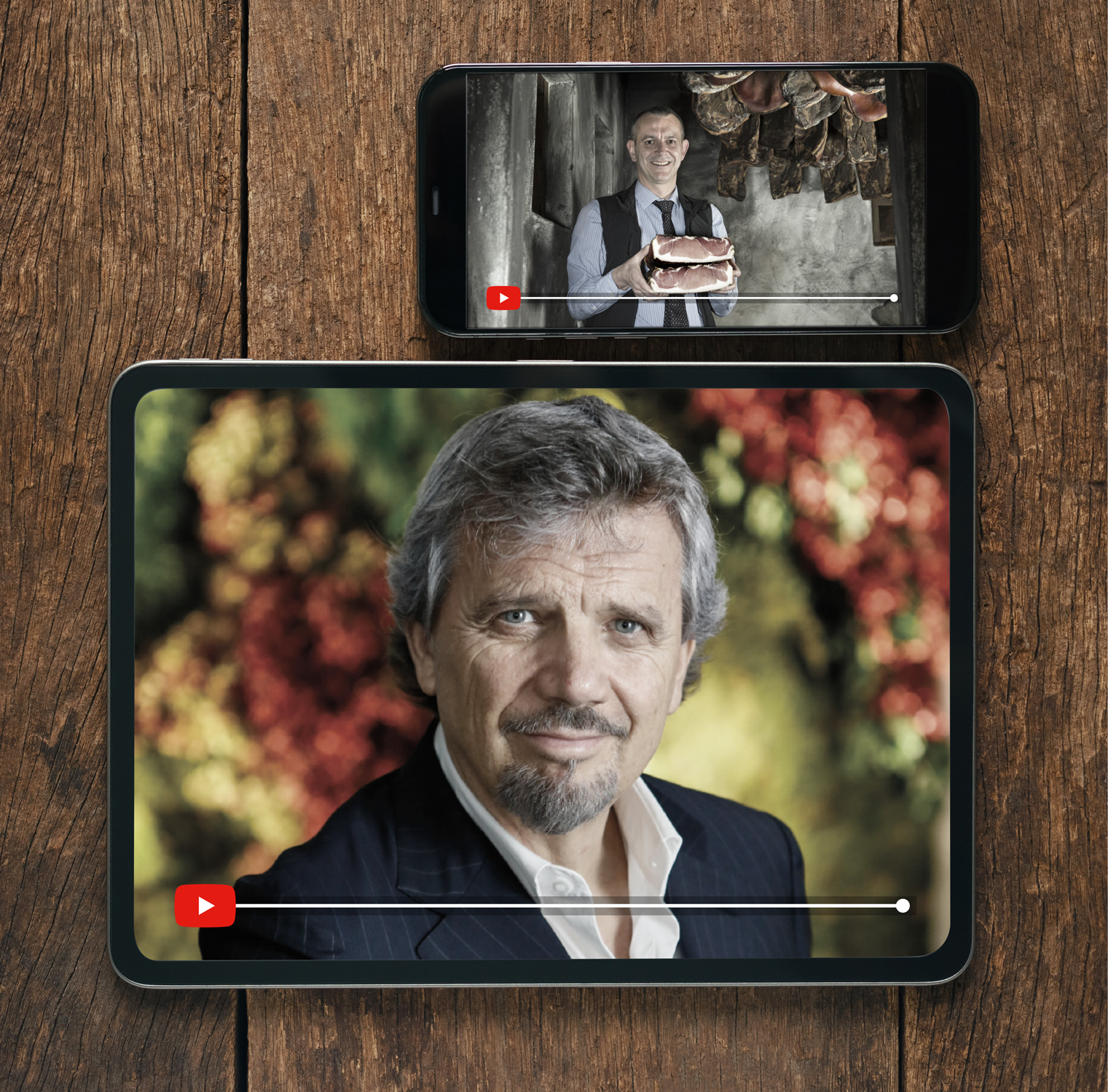 Un tablet e uno smartphone mostrano video che ritraggono due individui diversi. La tavoletta mostra un uomo anziano con i capelli grigi e la barba, che indossa un abito, con uno sfondo naturale sfocato. Lo smartphone mostra un uomo che tiene in mano un grosso pezzo di salume in un ambiente rustico.