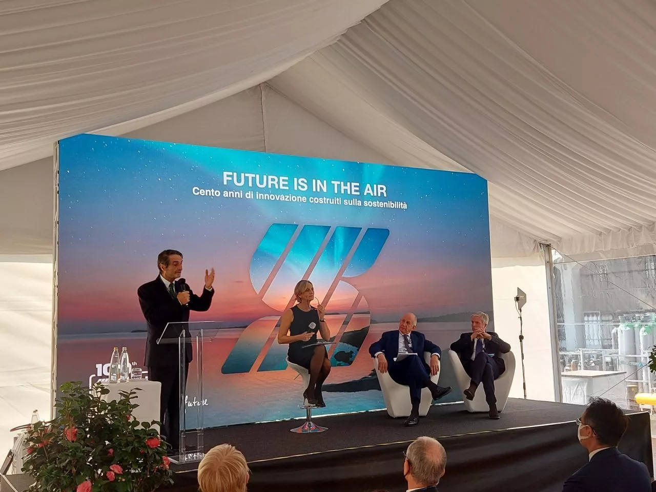 Un uomo parla su un podio sotto una tenda con uno striscione con la scritta "IL FUTURO È NELL'ARIA" e "Cento anni di innovazione realizzati sulla sostenibilità". Tre persone, due uomini e una donna, si siedono su un palco dietro di lui con sullo sfondo un tramonto e una scena sull'oceano.