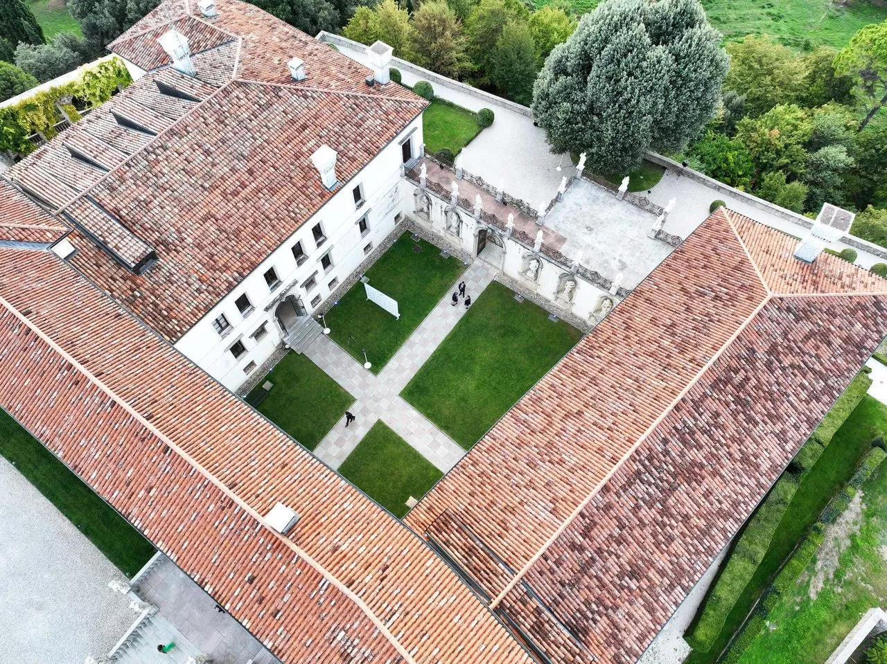 Veduta aerea di un edificio storico con tetti di tegole rosse che formano una forma a U attorno a un cortile centrale. Quattro persone camminano sui vialetti del cortile, fiancheggiato da prati verdi. Il verde circostante comprende grandi alberi e siepi ben curate.