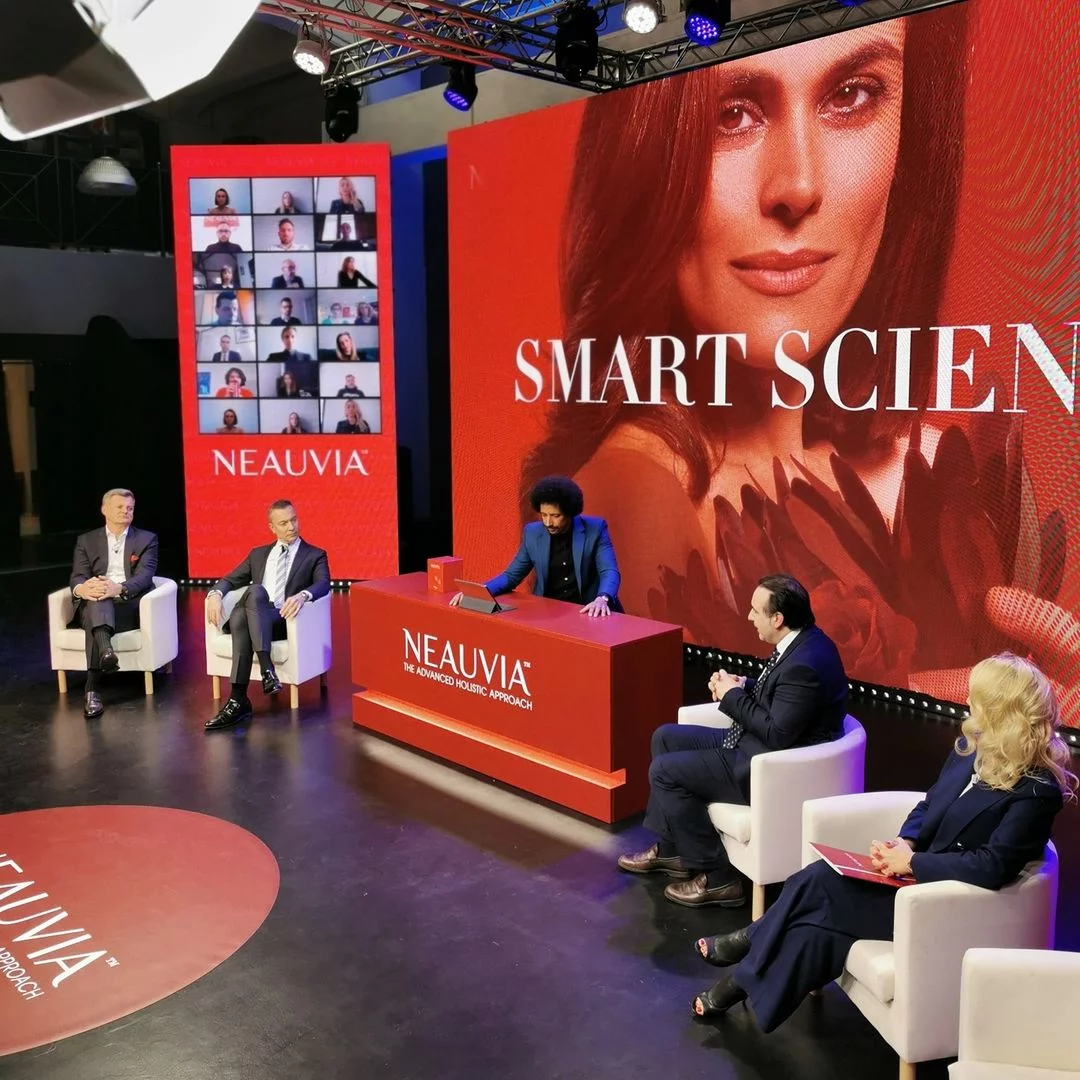 Una tavola rotonda si sta svolgendo in uno studio con quattro persone sedute, una su un podio. Dietro di loro c'è un grande schermo che mostra il marchio "NEAUVIA" e "SMART SCIENCE". Sullo schermo è in corso una riunione virtuale in cui vengono visualizzati diversi partecipanti.