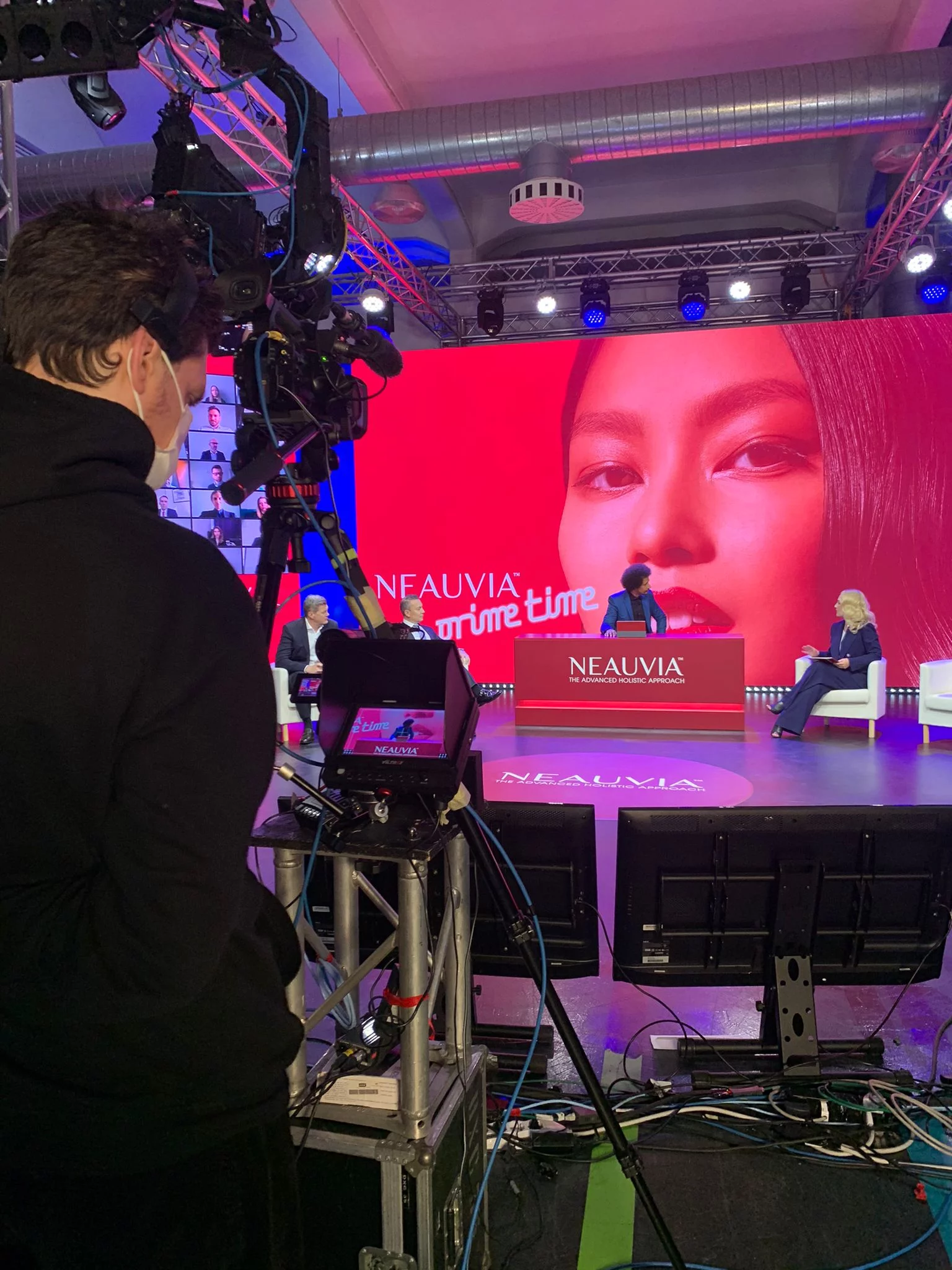 Veduta del dietro le quinte di un set; un operatore della telecamera sta filmando un gruppo di tre persone sedute sul palco. Lo sfondo del palco è un grande schermo che mostra il volto di una donna e il testo "NEAUVIA" e "Prime Time". Il set è dotato di illuminazione brillante e attrezzature tecniche.