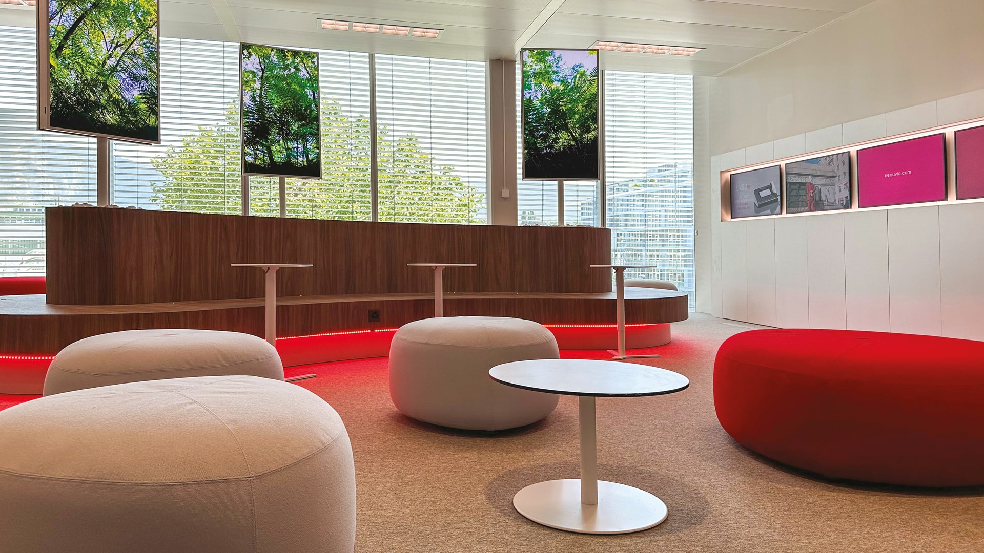 Modern lounge area with round white and red seating, small circular tables, and several screens displaying nature images. Large windows with blinds filter in natural light, creating a bright and inviting atmosphere.