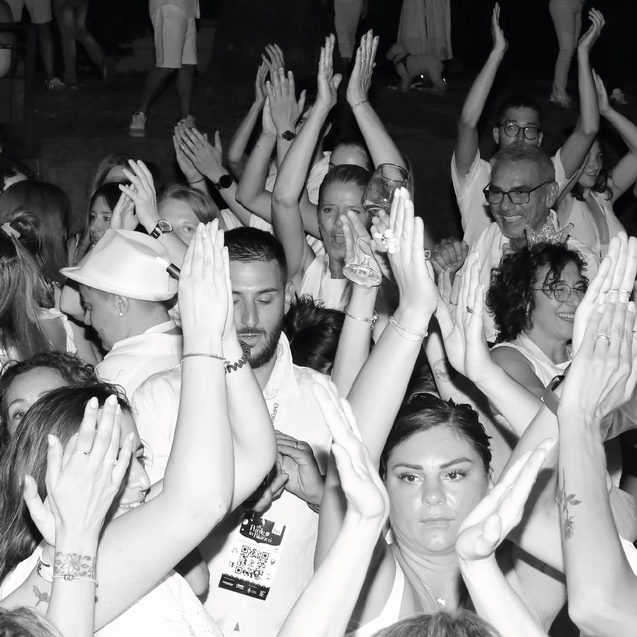 Un grande gruppo di persone vestite di bianco che applaudono con entusiasmo e alzano le mani in occasione di un evento o di una celebrazione di AI, con espressioni gioiose su molti volti. La foto è in bianco e nero.