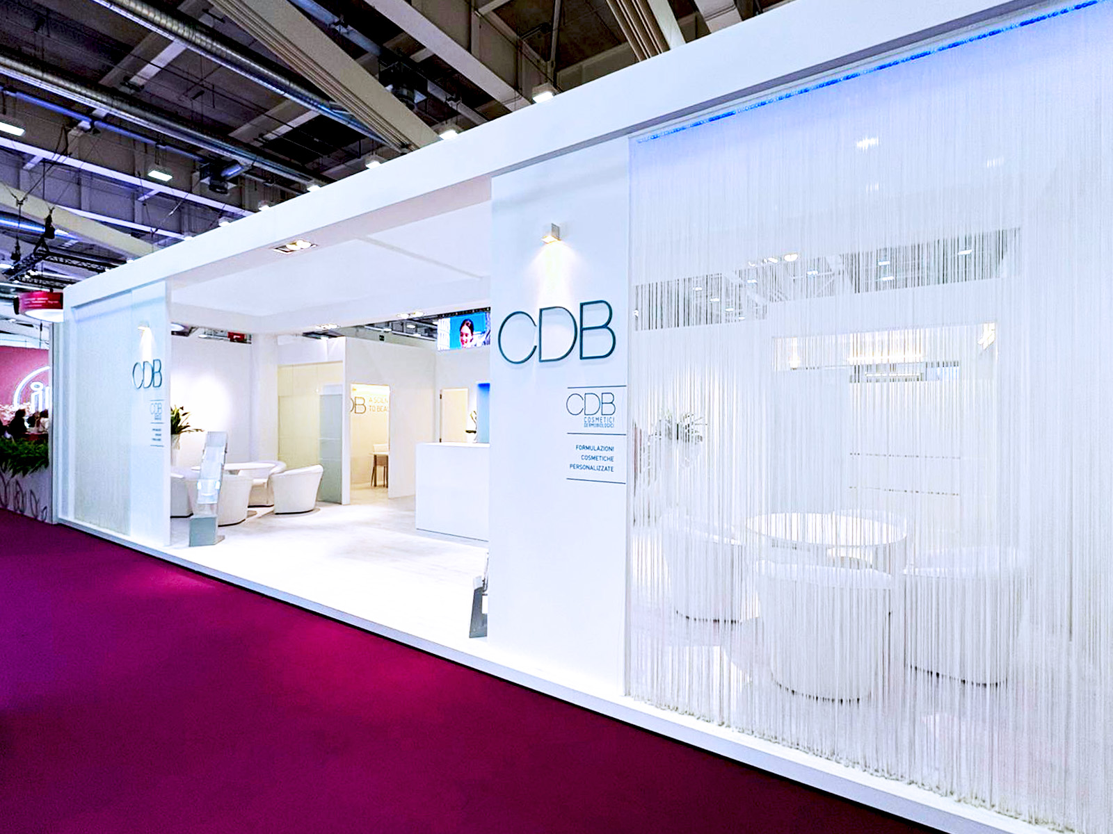Un moderno stand espositivo bianco con il marchio "CDB", caratterizzato da eleganti aree di seduta, tende con frange trasparenti e luci soffuse, su un pavimento in moquette magenta all'interno di una grande sala congressi.